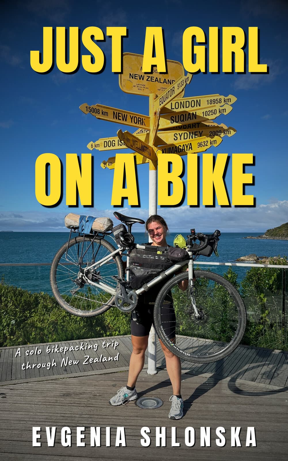Book cover: “Just a Girl on a Bike” by Evgenia Shlonska — Evgenia holding up her loaded bike beside a signpost above the sea