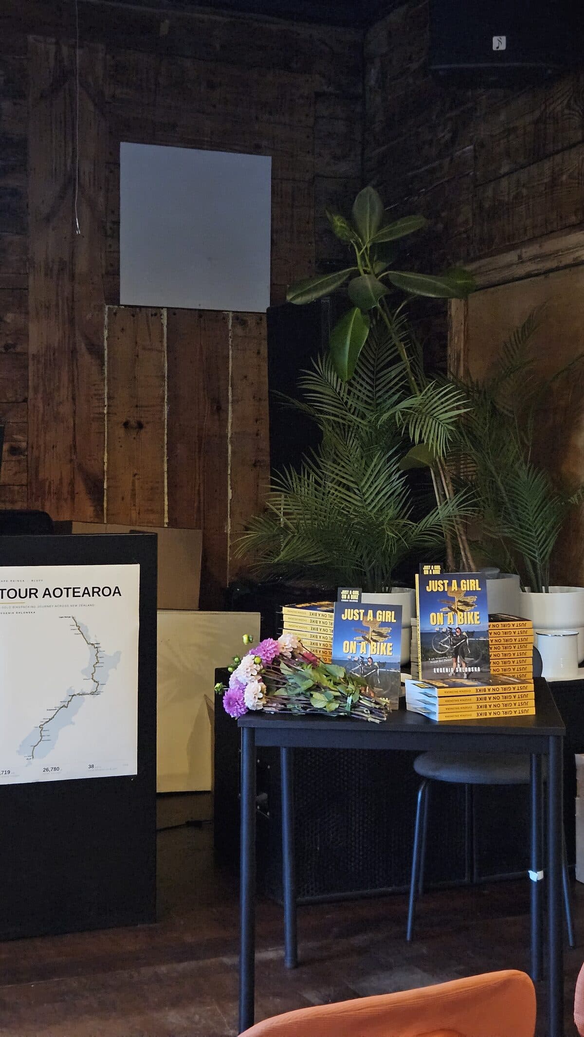 Stacks of the book on a table with flowers, next to a poster of the Tour Aotearoa route