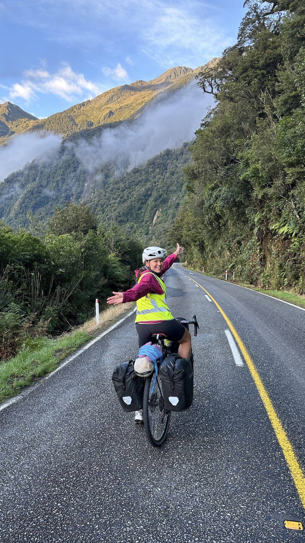 Evgenia riding a loaded bike on a mountain road in New Zealand, arms outstretched